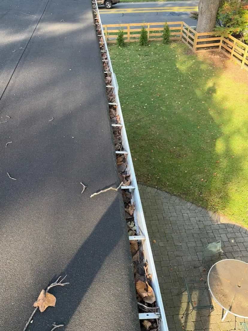 Gutter with leaves and twigs along a roof edge above a lawn and wooden fence