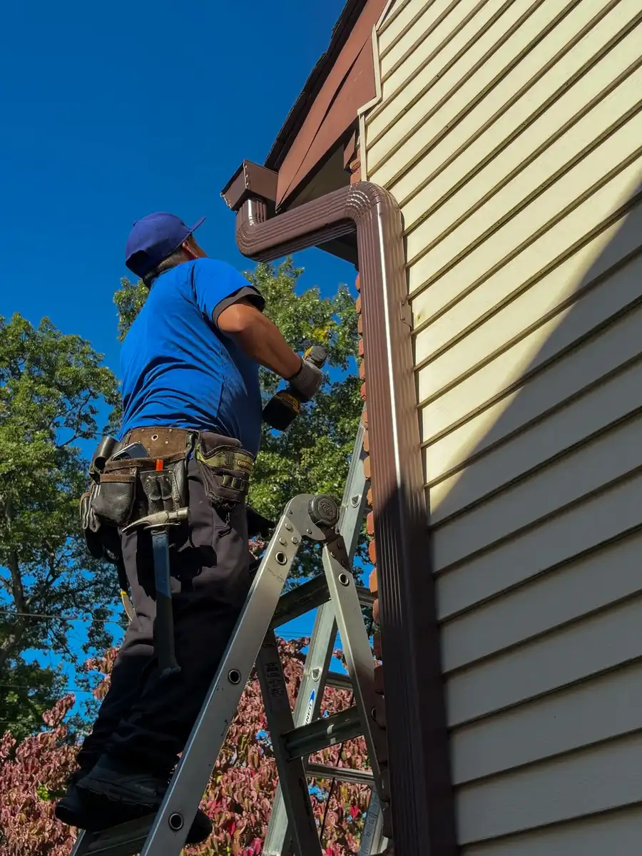 Tav crew member in a blue Tav shirt on a ladder fastening a new brown gutter and downspout at the roof edge, blue sky