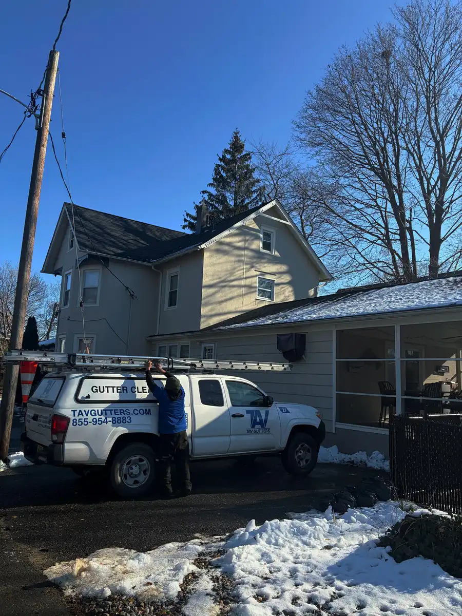 Tav Gutters pickup with ladders and a crew member in a blue Tav shirt beside it, parked in front of a house, blue sky