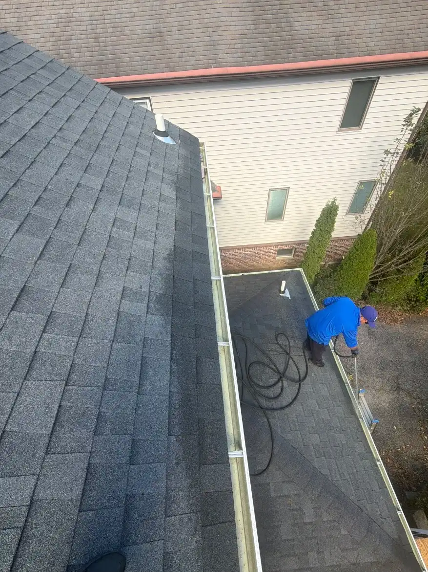 Crew member in a blue shirt working with a hose on a lower roof beside a gutter, seen from above