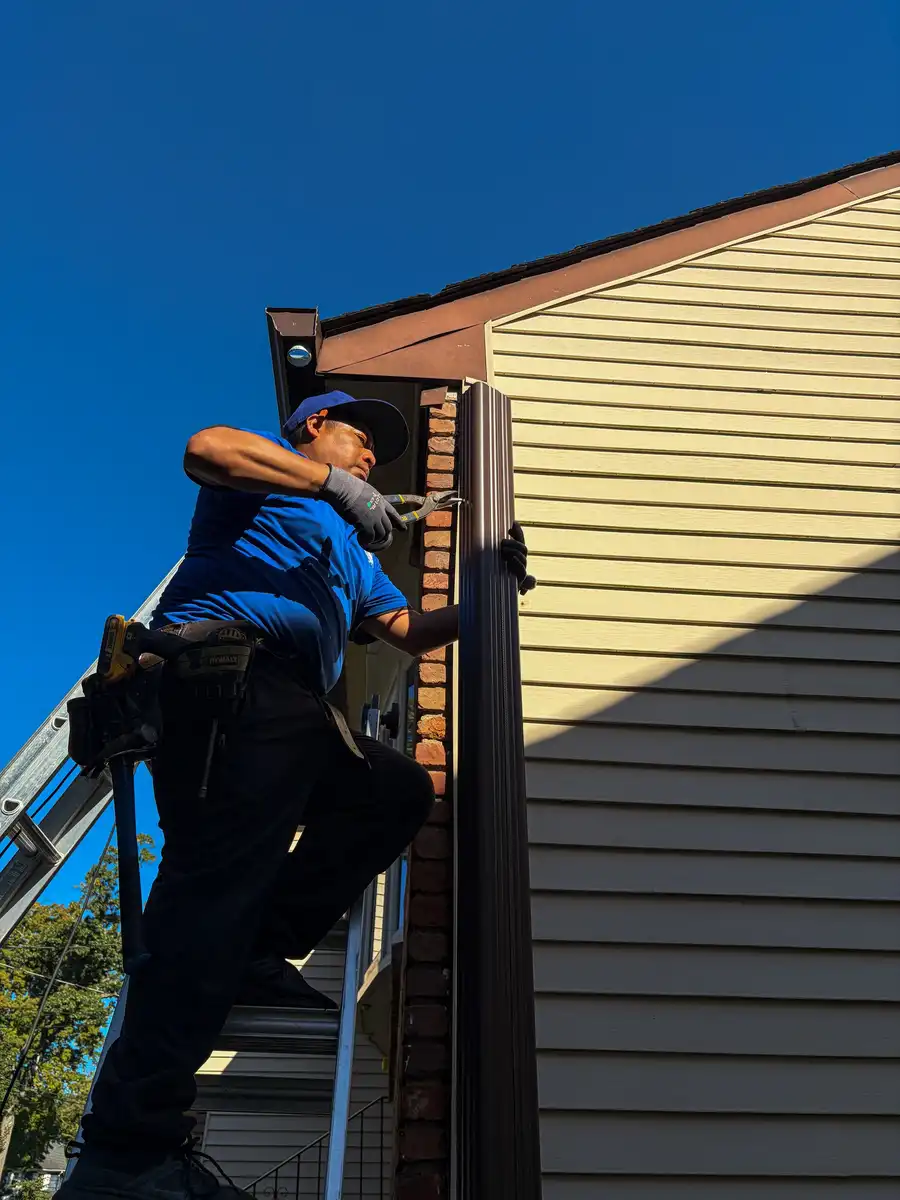 Tav crew member in a blue Tav shirt on a ladder working at the gutter corner of a brick and siding house, blue sky