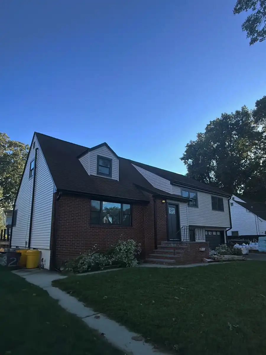 New black seamless gutters and downspout on a white-sided house with a dark roof and brick base, seen from the lawn