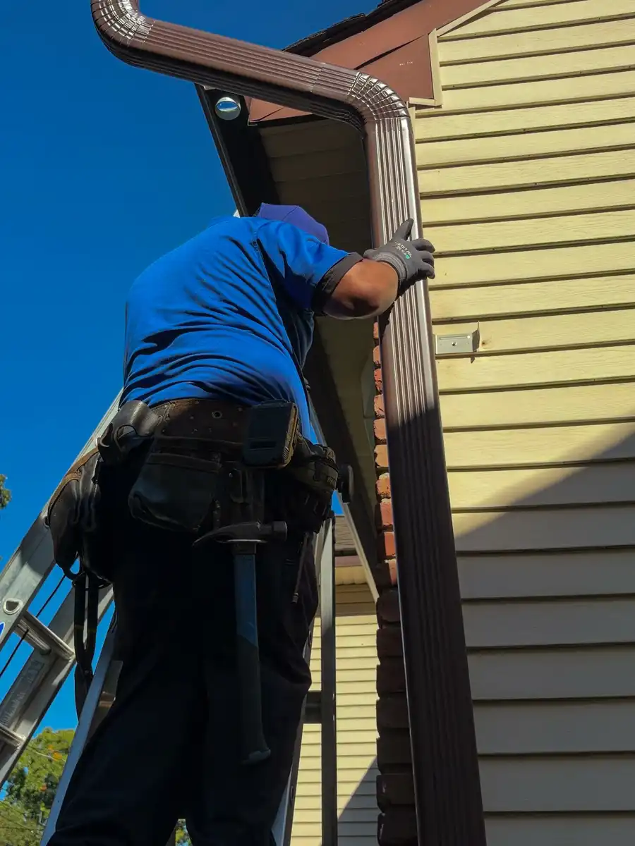 New brown gutter outlet and downspout elbow at the roof edge with the crew member fastening it from the ladder